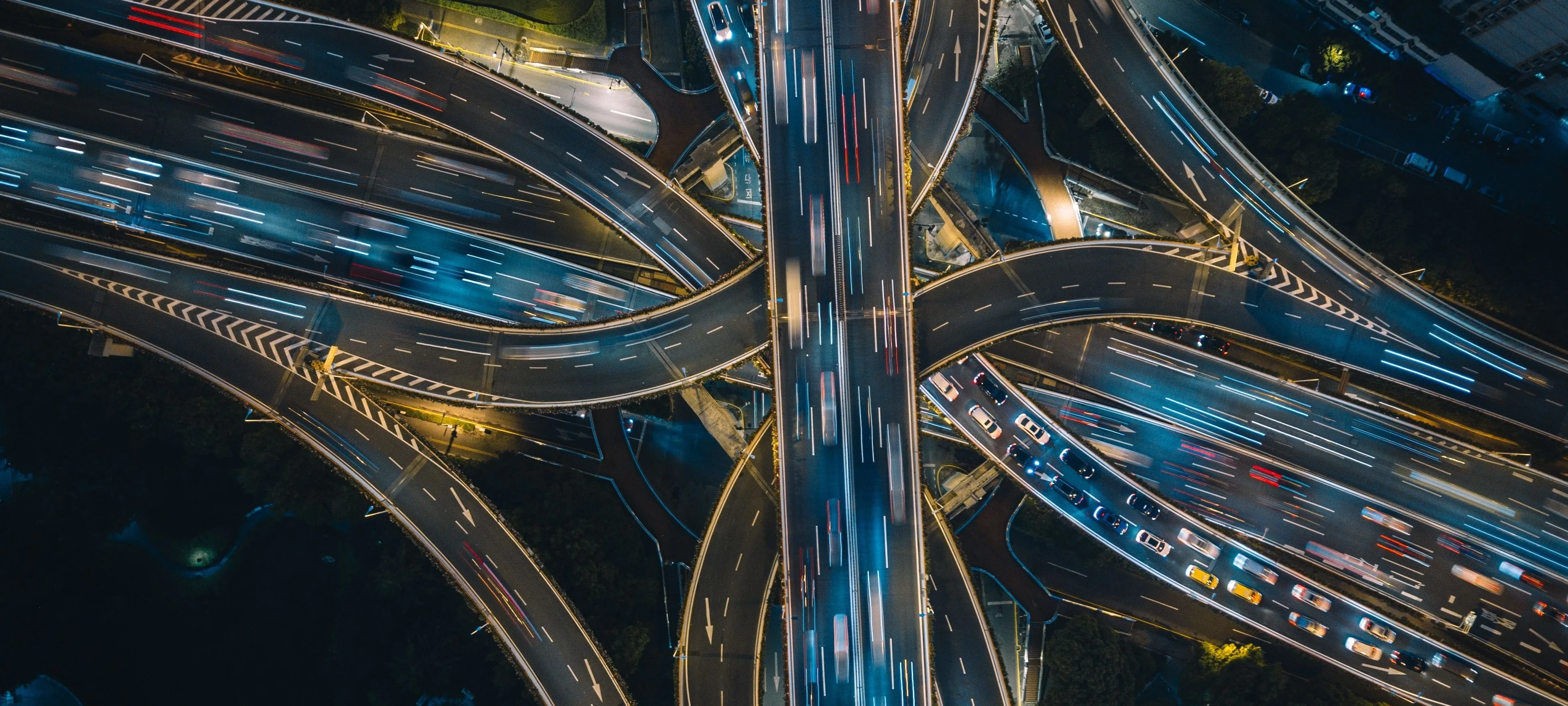 A city interchange from directly overhead at night, elevated roads crossing above lanes of moving traffic