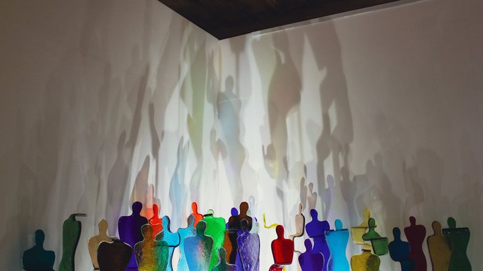A crowd of small coloured glass figures, their shadows cast large on the walls behind them