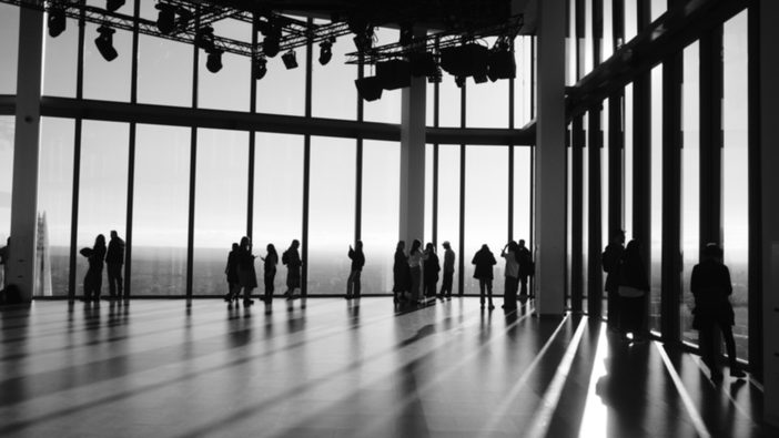 Figures silhouetted along a wall of glass on a high floor, sunlight striping the polished floor